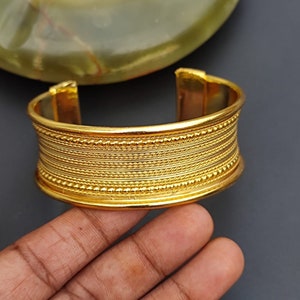 May include: A gold-toned metal cuff bracelet with a textured design. The bracelet has multiple rows of raised lines and small beads.