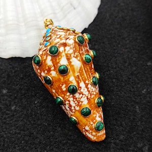 Könnte beinhalten: Ein dekorativer Muschelanhänger mit goldfarbener Öse und Akzenten. Der Anhänger ist mit grünen und türkisfarbenen Edelsteinen besetzt, die in goldfarbenen Fassungen gefasst sind. Die Muschel hat ein braun-weißes, gesprenkeltes Muster.