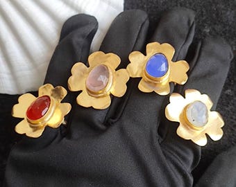 May include: Four gold-colored flower-shaped rings with different colored gemstones: red, pink, blue, and white. The rings are displayed on a black surface, with a white seashell in the background. The rings are likely fashion accessories.