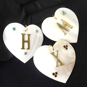 Op de afbeelding: Drie hartvormige hangers van wit parelmoer. Elke hanger heeft een gouden initiaal: H, M en X. De hangers zijn versierd met kleine, gekleurde edelstenen en gouden lussen om op te hangen.