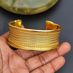 May include: A gold-toned bangle bracelet with a textured design. The bracelet has multiple rows of raised lines and small beads.