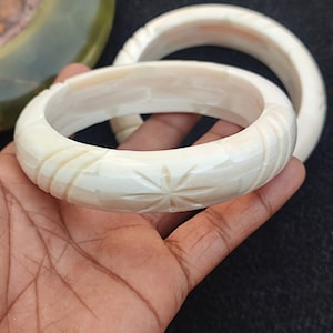 May include: Two white carved bone bangles. The larger bangle has a starburst design carved into the surface.