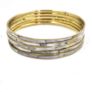 May include: A stack of six gold-toned bangles. Each bangle has a band of white and gray iridescent material. The bangles are stacked together, creating a layered effect. The background is plain white.