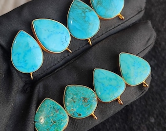 May include: Six turquoise gemstone earrings with gold-colored settings. The teardrop-shaped stones vary in shades of blue and green. The earrings are displayed on a black surface, with a seashell in the background.