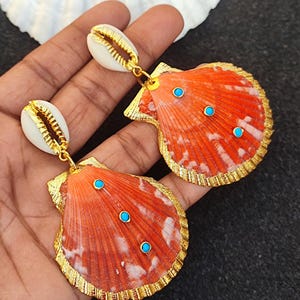 Puede incluir: Un par de pendientes de concha naranja con detalles dorados y piedras turquesa. Los pendientes son colgantes y tienen una concha blanca más pequeña en la parte superior.