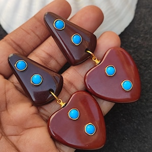May include: A pair of earrings with a geometric design. The earrings are made of brown and red resin and feature turquoise stones. The earrings are dangle style and have a heart-shaped pendant.