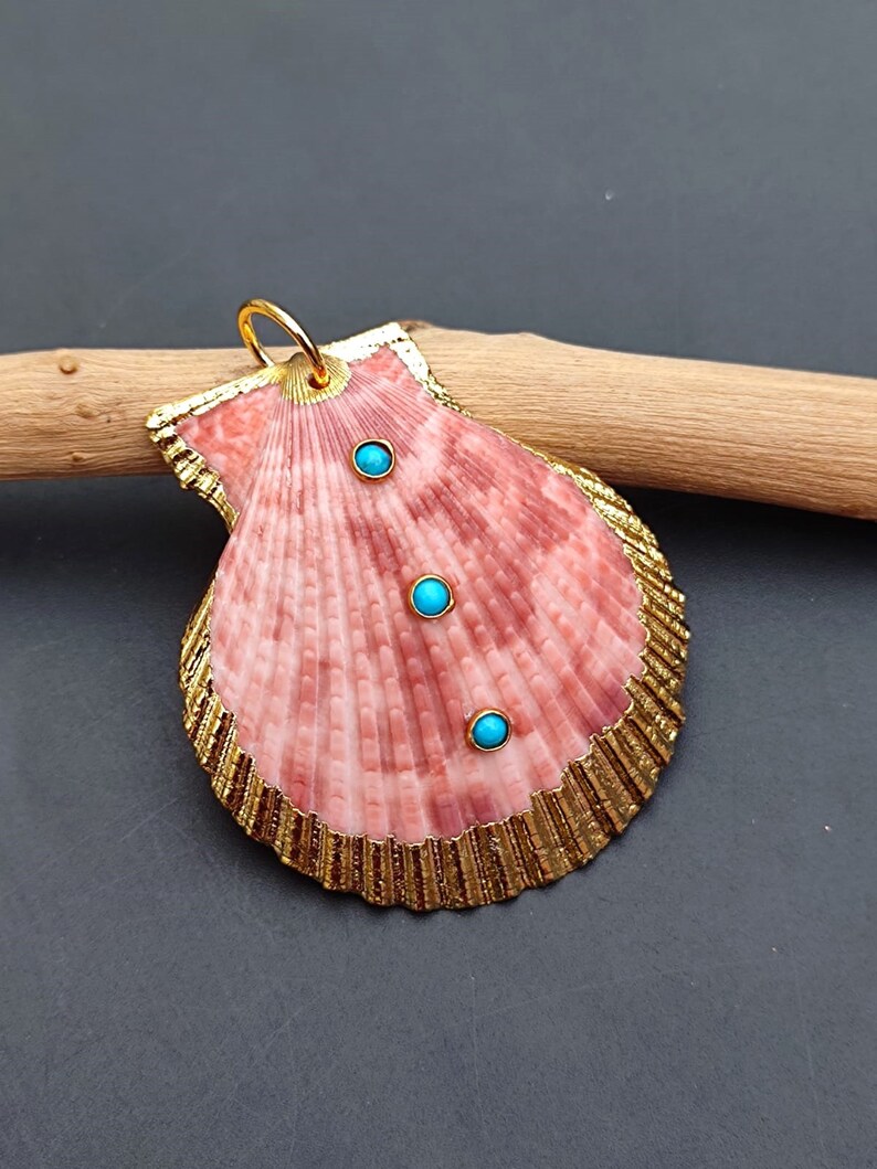 K&ouml;nnte beinhalten: Ein rosa Muschel-Anh&auml;nger mit goldener Umrandung und drei t&uuml;rkisfarbenen Steinen. Der Anh&auml;nger h&auml;ngt an einer goldenen Kette.