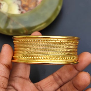May include: A gold-toned bangle bracelet with multiple rows of decorative detailing. The bracelet has a smooth, polished finish.