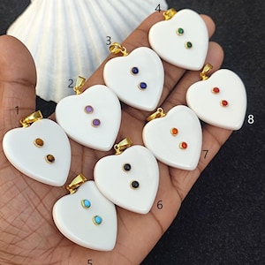 May include: Eight white heart-shaped pendants with gold bail and two gemstone accents. The gemstones are different colors, including blue, green, red, orange, purple, black, and turquoise.