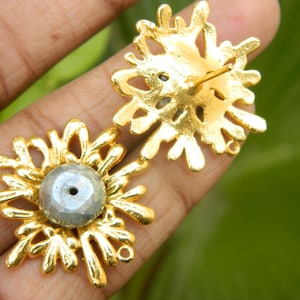 May include: A pair of gold earrings with a grey gemstone in the centre of each earring. The earrings are shaped like a flower with many petals.