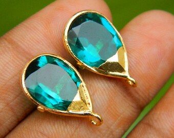 May include: Two teardrop-shaped teal gemstones with gold-colored settings. The gems have a faceted cut, reflecting light. Each setting has a small loop at the bottom. The background is a blurred green.