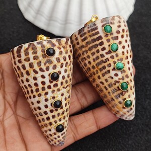 May include: Two seashell pendants with gold-colored bails. One pendant has three black stones, the other has three green stones. The shells have a natural, speckled pattern. The pendants are held in a hand against a dark background.