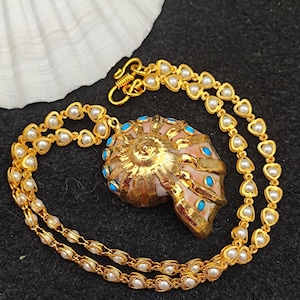 Könnte beinhalten: Eine goldfarbene Muschel-Anhänger-Halskette mit einer Perlen- und herzförmigen Kette. Die Muschel ist in Rosa- und Goldtönen bemalt und mit türkisfarbenen Steinen verziert. Die Halskette wird auf einer schwarzen Oberfläche präsentiert.