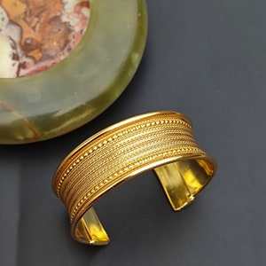 May include: A gold-toned metal cuff bracelet with a textured design. The bracelet has a wide band with multiple rows of raised lines and small beads.
