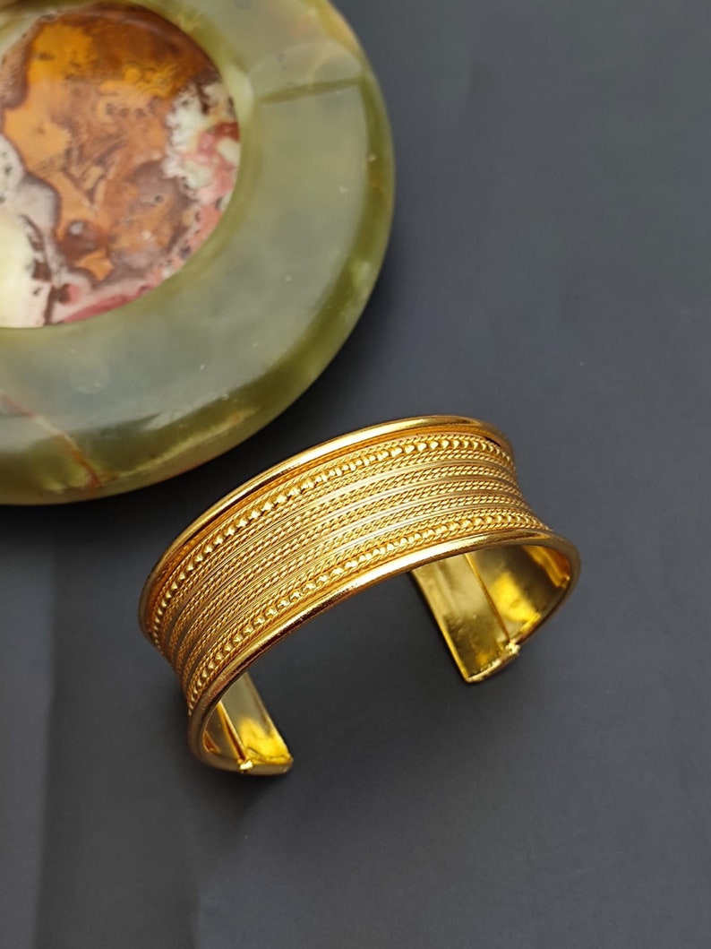 May include: A gold-toned metal cuff bracelet with a textured design. The bracelet has a wide band with multiple rows of raised lines and small beads.
