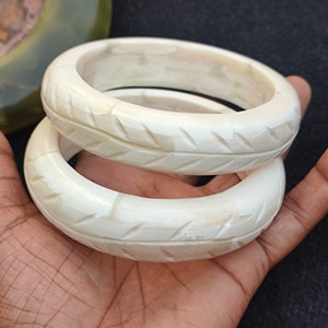 May include: Two ivory-coloured bangles with carved diagonal lines. The thick, round bracelets are stacked on top of each other, showcasing their detailed design. The bangles are displayed against a dark background.