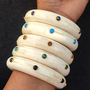 May include: A set of six ivory bangles with gold accents and colourful gemstones. The bangles are stacked on a person's wrist.
