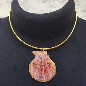 Peut inclure: Un collier ras du cou doré avec un pendentif en forme de coquillage rose bordé d'or. Le pendentif est orné de trois pierres turquoise et de deux pierres marron. Le collier est présenté sur un mannequin noir.
