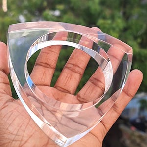 Op de afbeelding: Transparante acryl armband met een driehoekige vorm. De transparante armband heeft een glad, gepolijst oppervlak en een cirkelvormige opening in het midden. De armband wordt in een hand gehouden tegen een vage groene achtergrond.