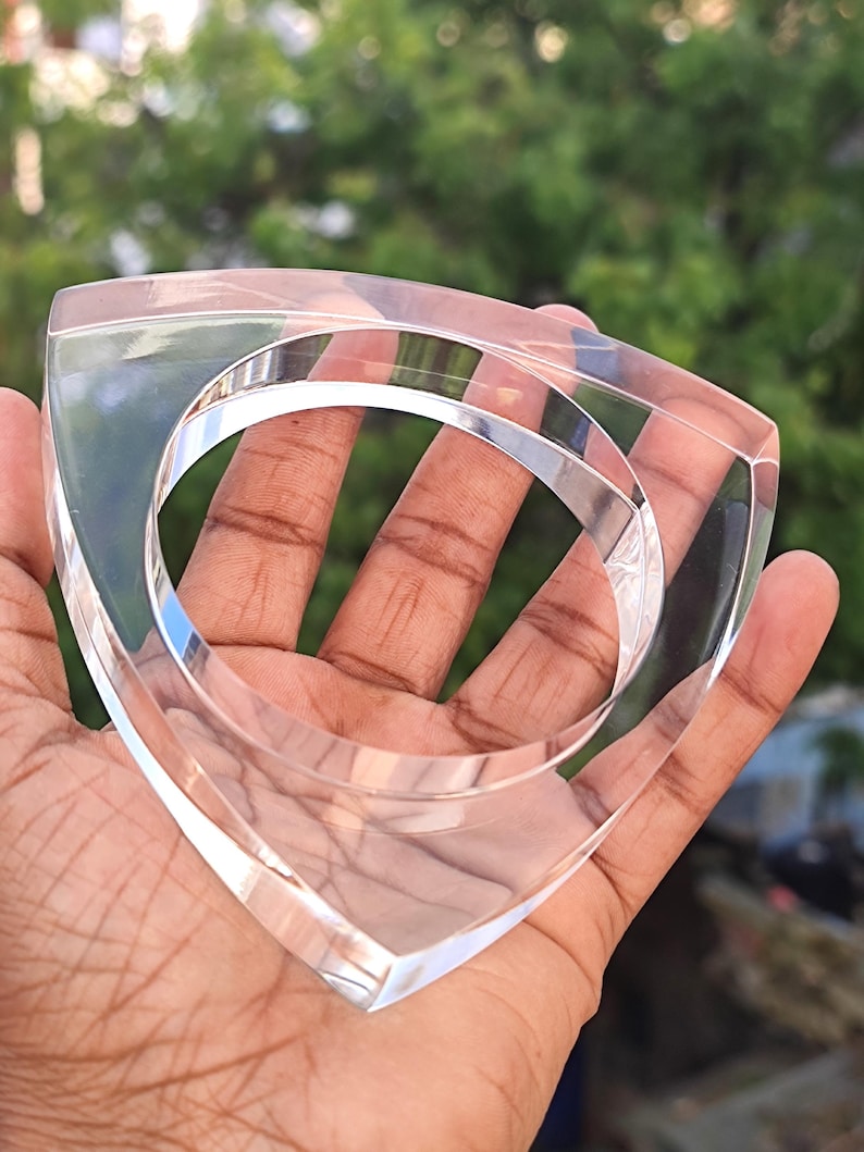 Op de afbeelding: Transparante acryl armband met een driehoekige vorm. De transparante armband heeft een glad, gepolijst oppervlak en een cirkelvormige opening in het midden. De armband wordt in een hand gehouden tegen een vage groene achtergrond.