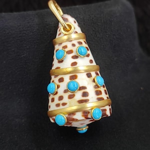Peut inclure: Pendentif en forme de cône, composé d'une coquille tachetée de brun, ornée de bandes dorées et d'accents turquoise. Le pendentif est doté d'une bélière dorée pour la suspension. La coquille mesure environ 3,8 cm de long.