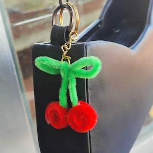 Puede incluir: Un llavero de cereza roja y verde colgando de un bolso negro. Las cerezas son esponjosas y las hojas son verdes. El llavero está sujeto a un anillo dorado y una correa negra.