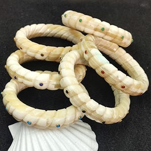 May include: A collection of cream-coloured, carved bracelets with colourful gemstone accents. The bracelets are stacked on a dark surface, with a white seashell in the foreground. The bracelets have a textured, rope-like design.