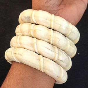 May include: A set of five ivory-coloured bangles with a twisted design. The bangles are stacked on a person's wrist.