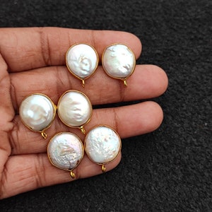 Könnte beinhalten: Fünf weiße Perlencabochons mit goldfarbenen Fassungen und Ösen. Die Perlen sind rund und haben eine leicht unregelmäßige Form. Die Perlen sind in ein goldfarbenes Metall gefasst.