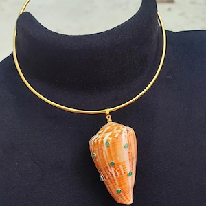 May include: A gold-colored choker necklace with a pendant featuring a large, orange and white seashell. The shell is adorned with small, green embellishments. The necklace is displayed on a black surface.