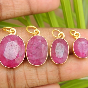 May include: Four oval-shaped ruby pendants with gold-colored settings. The rubies are a deep pink color and have a faceted cut. Each pendant has a small loop at the top for attaching to a chain or other jewelry piece.