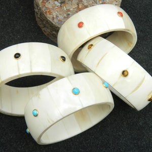 May include: Four ivory bangles with different colored gemstones. The bangles are stacked on top of each other. The gemstones are turquoise, black, orange, and tiger's eye.