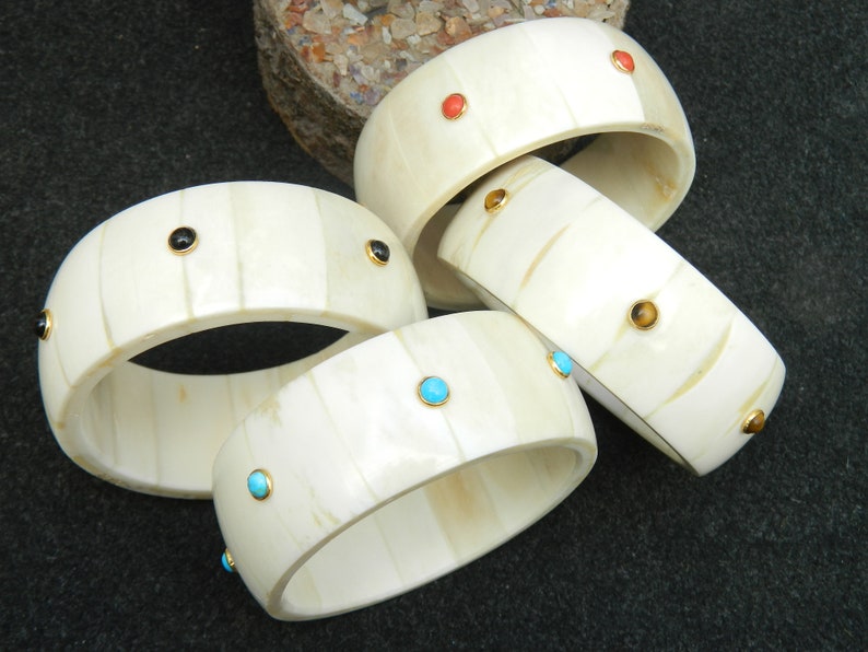 May include: Four ivory bangles with different colored gemstones. The bangles are stacked on top of each other. The gemstones are turquoise, black, orange, and tiger's eye.