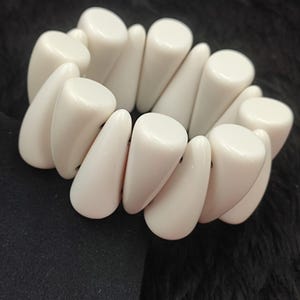 May include: Ivory-colored bracelet with a chunky, geometric design. The bracelet features rounded cylindrical and teardrop-shaped beads, creating a bold, statement piece. The beads are strung together to form a flexible, wearable accessory.