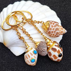 Pode incluir: Quatro chaveiros de concha do mar banhados a ouro com detalhes turquesa e pretos. Os chaveiros estão presos a uma corrente dourada e um conjunto de argolas de chaveiro.