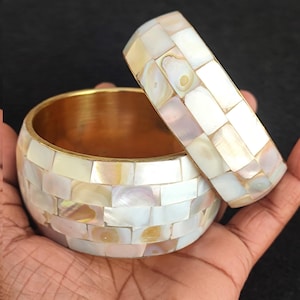 Puede incluir: Dos pulseras blancas y doradas con un diseño de mosaico hecho de pequeñas piezas de concha iridiscente.