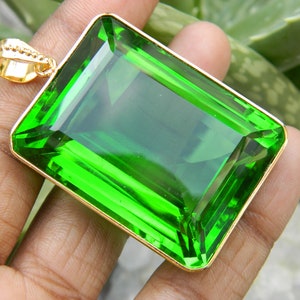Puede incluir: Un colgante de piedra preciosa de color verde esmeralda grande con un engaste de oro. El colgante tiene forma rectangular y está tallado a facetas.