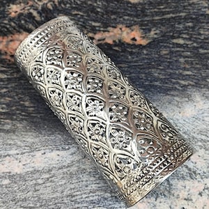May include: Silver metal cuff bracelet with an intricate, ornate design. The cuff is cylindrical and has a textured surface.