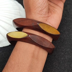 May include: Two wooden bangles with gold accents. The bangles are brown and have a smooth, polished finish. The gold accents are in the shape of ovals and are located on the top and bottom of the bangles.