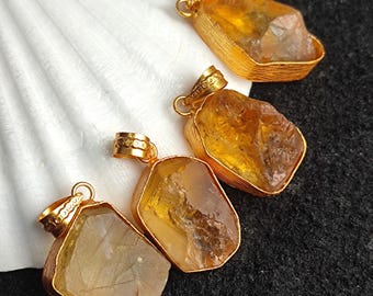 Puede incluir: Cuatro colgantes de citrino en bruto con biseles dorados. Las piedras de citrino, de formas variadas, presentan un cálido tono amarillo dorado. Los colgantes se exhiben sobre una concha blanca y una superficie gris oscuro.