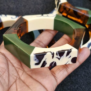 May include: A set of four hexagonal bangle bracelets. The bracelets are made of a multi-colored plastic material. The colors are green, brown, white, and black.