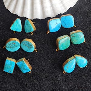 May include: A collection of turquoise stud earrings with gold-colored metal settings. The earrings feature various shapes, including triangles, squares, and teardrops. A white seashell is in the background, and the earrings are displayed on a dark gray surface.