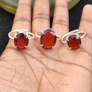 May include: Three silver rings with oval-shaped red gemstones. The rings are on a person's hand.