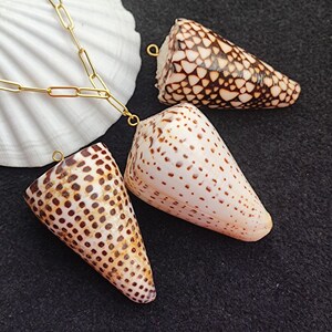 May include: Three cone-shaped seashells with brown and white patterns, each with a small gold loop attached. A gold chain is connected to one shell, and a large white seashell is in the background. The shells are on a black surface.