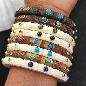 May include: A stack of wooden and bone bangles featuring turquoise, green, and blue gemstones. The bangles are carved with intricate designs and have gold accents.