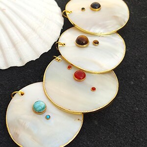 May include: Four white, oval-shaped pendants with gold trim and various gemstone accents, including turquoise, red, and brown stones. A white seashell is visible in the background. The pendants are likely jewelry.
