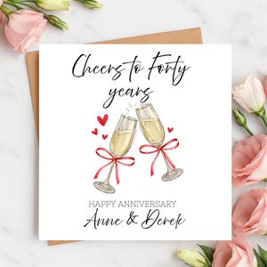 Può includere: Biglietto di anniversario bianco con la scritta "Cheers to Forty years" e "Happy Anniversary Anne & Derek". Due calici di champagne con fiocchi rossi si toccano, circondati da cuori rossi.