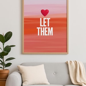 May include: A framed art print with a red heart above the white text "LET THEM" against a gradient background of pink, orange, and red. The artwork is displayed above a gray sofa with a cream-colored pillow and blanket.