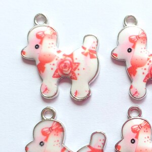May include: Four silver charms shaped like poodles with a pink and white floral design. The charms have a loop at the top for attaching to a necklace or bracelet.
