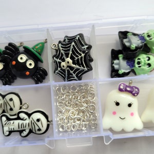 May include: A clear plastic storage box with compartments filled with Halloween-themed charms. The charms include spiders, ghosts, skeletons, and a witch's hat. There are also silver jump rings and a spiderweb charm.